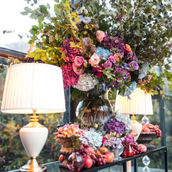 Console table topped with wild florals, grapes, and glass lamps in a sunlit event space